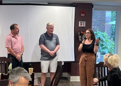Two PTP team members stand alongside a woman holding a microphone, speaking to guests at Hope Lodge Boston during a community dinner event, with a presentation screen in the background.