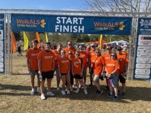 A group of PTP employees and their family members wearing orange WalkALS shirts, standing together at the starting line of the ALS Walk in Cincinnati under a banner that reads 